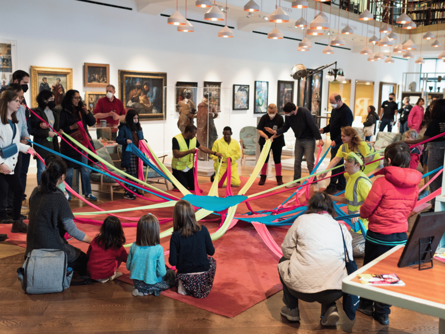 A group of people, including BLINK facilitators wearing high-vis tops, are dancing with an octoband; a brightly-coloured lycra circle with long arms coming off that everyone can hold on to