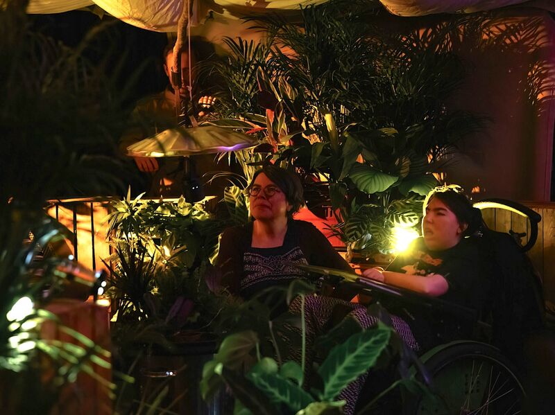 A young woman using a wheelchair and a middle-aged woman sit  beside each other. They are surrounded by wooden crates and plants, with a billowing tent canopy overhead. Atmospheric low lighting, illuminates their faces with a golden glow and casts shadows of the plant leaves around them.