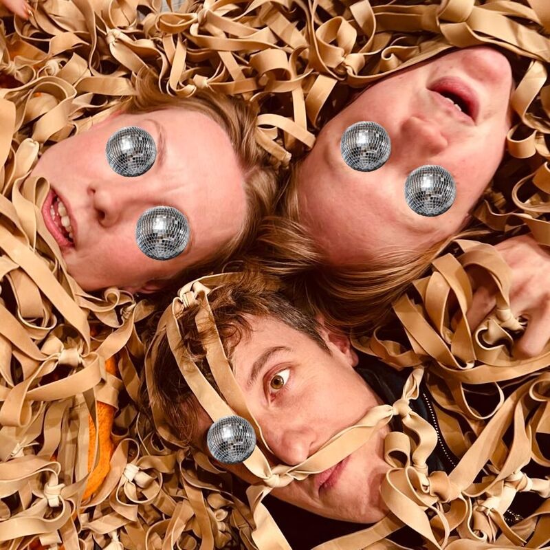 SKSie with their eyes covered with glitter balls and faces emerging from packing paper strips