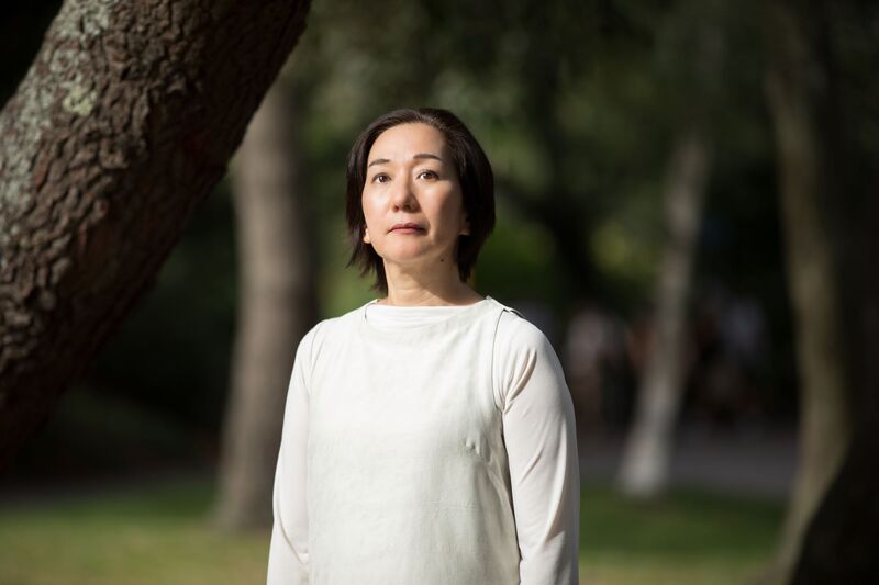 Chisato, a Japanese woman with a black bob. She stands in a white tunic dress in front of a blurred background of trees and greenery.