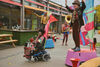 Black woman in wheelchair with colourful flag attached drives past two men in colourful clothes, stood on coloured blocks with bright flags on