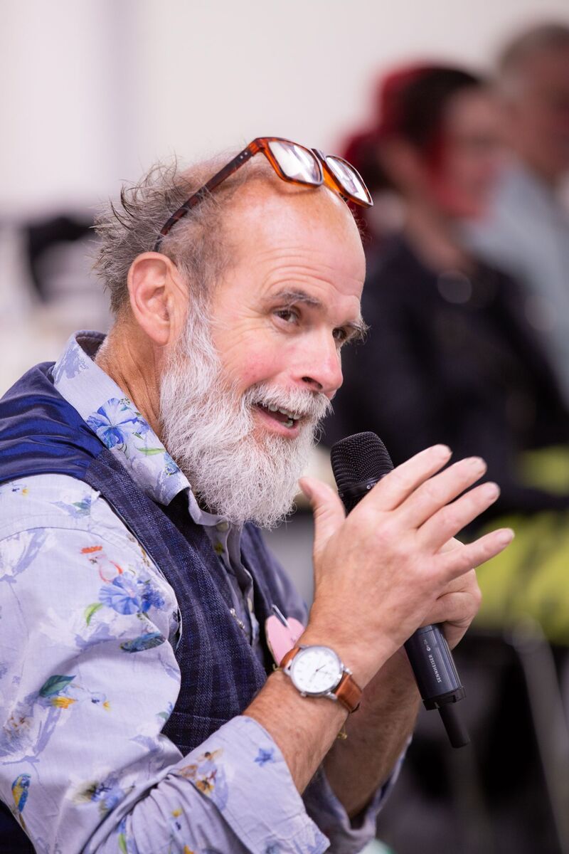 Lol O'Connor, who has a full, white beard, bald on most of the top of his head and curly grey-white hair around the remainder of his head in three-quarter profile, holding a microphone, reading glasses perched on his forehead, smiling, with one hand gesturing,  open