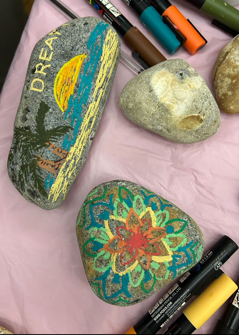 Two pebbles painted by hand, one with a flower image in rainbow colours and the other an image of a palm tree and sunset and hte word 'DREAM' painted on it.