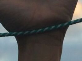 A photograph of an outstretched hand reaching up towards the sky, with a taut green rope running horizontally across the wrist. The background features a row of residential buildings with lit windows at dusk, under a sky with soft clouds illuminated by the setting sun.