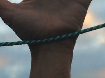 A photograph of an outstretched hand reaching up towards the sky, with a taut green rope running horizontally across the wrist. The background features a row of residential buildings with lit windows at dusk, under a sky with soft clouds illuminated by the setting sun.