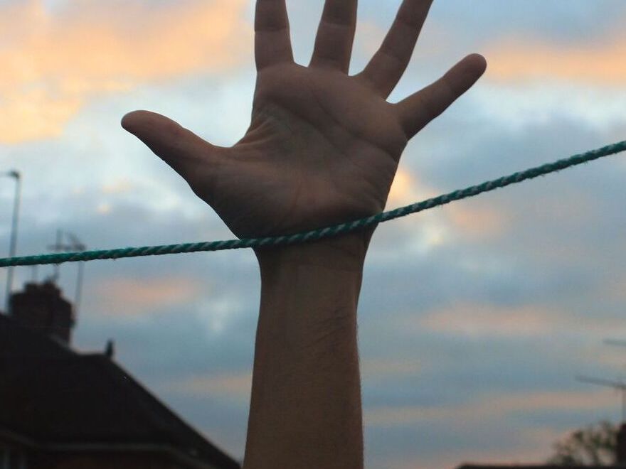 A photograph of an outstretched hand reaching up towards the sky, with a taut green rope running horizontally across the wrist. The background features a row of residential buildings with lit windows at dusk, under a sky with soft clouds illuminated by the setting sun.