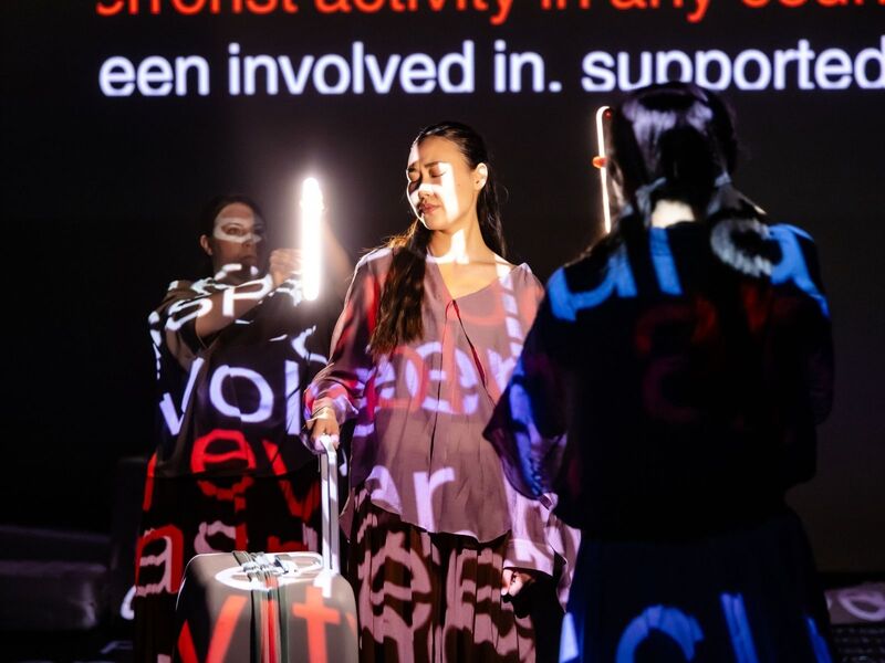Production shots from Transit - the first is a group of actors crowded around a tower of suitcases while one pretends to take a selfie using a smaller suitcase; the second is a group of actors standing behind a row of suitcases with projections describing how much they've spent on migrating to the UK; the third is a group of actors clumped together in low light, their arms stretched out to the ceiling mid-contemporary dance; the fourth is an actor standing in between two harsh lights with words projected over her face; the fifth is five actors standing in the corners and centre of the stage, 'talking' to the home office via mimed phones.