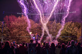 Firework flares rising into a dark sky, silhouettes of people's heads below, trees behind the fireworks