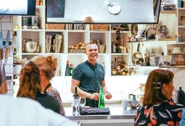 A White Man Wearing A Forrest Green T-Shirt  With Short Hair Smiles Whilst Giving A Demonstration To A Bustling Room Of People