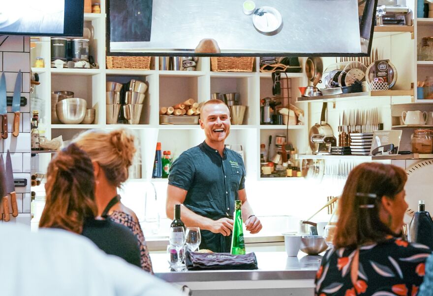 A White Man Wearing A Forrest Green T-Shirt With Short Hair Smiles Whilst Giving A Demonstration To A Bustling Room Of People