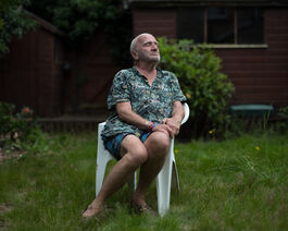 An older gentleman sits on a white plastic chair in a garden, with his eyes closed