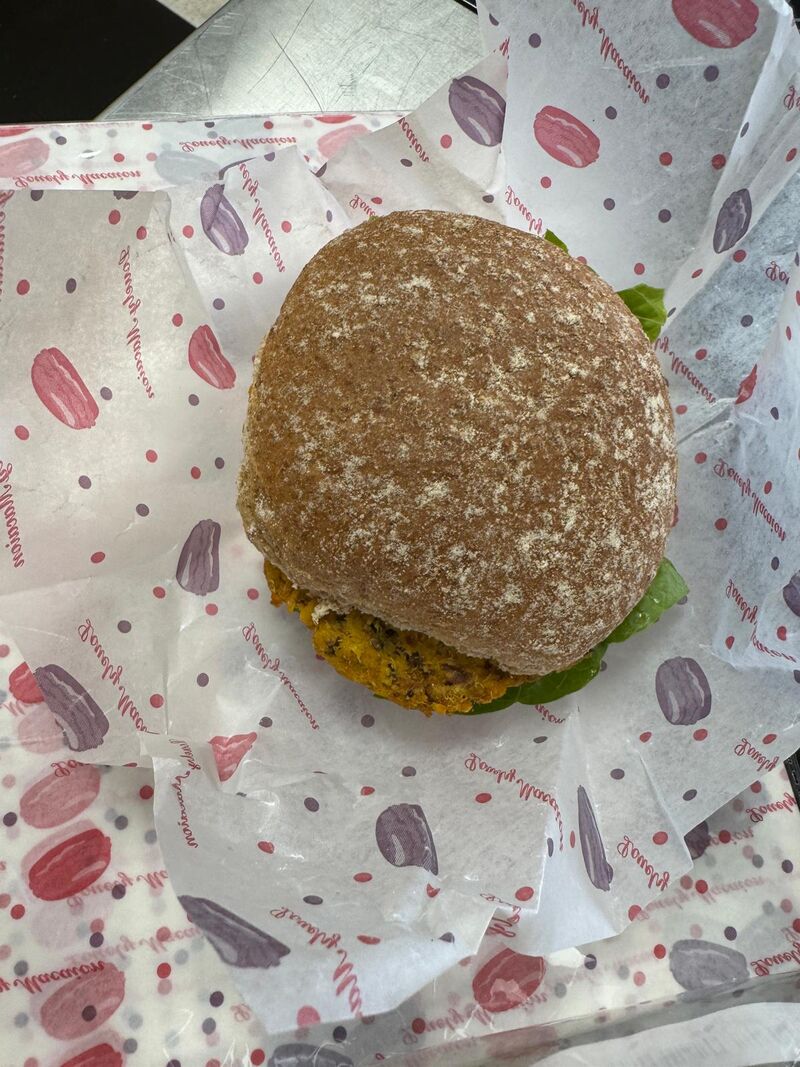 A delicous -looking whole homemade burger bun, sits on colourful grease proof paper. A burger and salad is visible from under the bun.