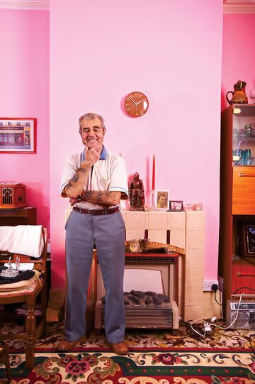 An older man smiling proudly in a home with pink walls and vintage decoration.
