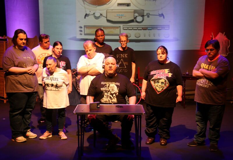 Everyone in the group is dressed in t-shirts featuring iconic music artists or bands, including The Beatles, Elton John, Queen, Guns N’ Roses, Iron Maiden, and Pink Floyd. Behind them, a large image of a vintage TEAC reel-to-reel tape recorder is projected onto a screen. The stage lighting is moody with purple and red hues, adding to the dramatic tone of the scene.