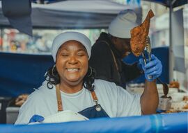 A Black Woman is smiling and holding up a piece of Jerk Chicken