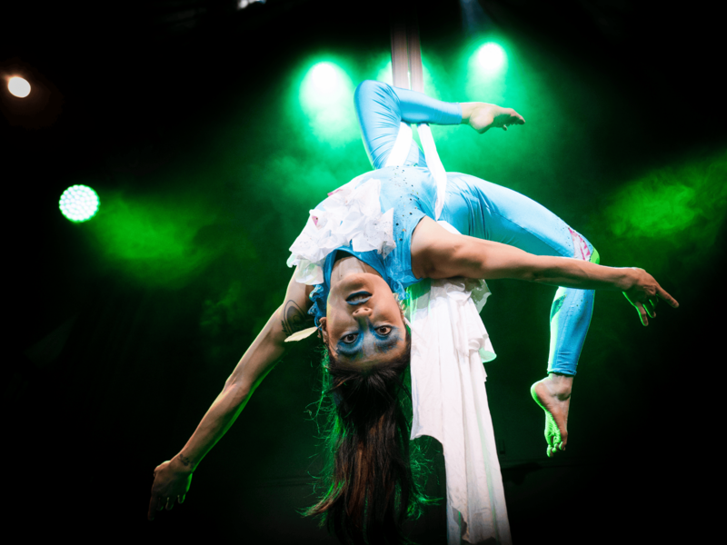 A performer in a blue costume suspended upside down on white aerial silks, with green stage lighting in the background