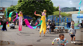 Children play in a playground while two women on stilts in coloursful clothing walk through.