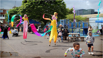 Children play in a playground while two women on stilts in coloursful clothing walk through.