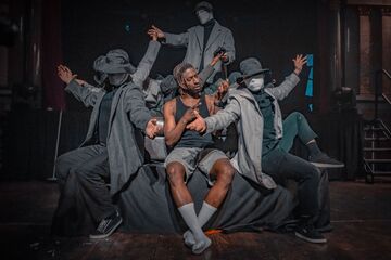 Sim Campbell sits centre stage on a sofa. He is a black man with dyed dreadlocks, wearing a black vest, grey shorts and white socks. He's surrounded by mysterious figures wearing trench coats, masks and hats. They frame him as they pass objects to eachother, behind and around him. But Sim is looking intently at his own hands, which are held up in front of him as he uses sign language.