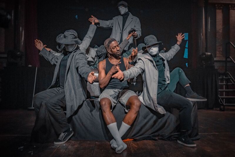 Sim Campbell sits centre stage on a sofa. He is a black man with dyed dreadlocks, wearing a black vest, grey shorts and white socks. He's surrounded by mysterious figures wearing trench coats, masks and hats. They frame him as they pass objects to eachother, behind and around him. But Sim is looking intently at his own hands, which are held up in front of him as he uses sign language.