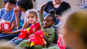 A crowd of children, focused on two girls, one of which has her hand up ready to speak.
