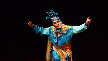 A man wearing an intricate blue and gold costume poses with his arms out and curved up.