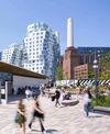A bustling urban plaza with modern, white curved buildings and iconic chimneys in the background. People stroll and relax under a clear blue sky.