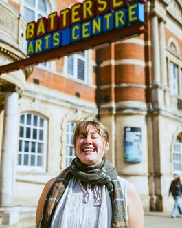 White woman wearing grey vest and dark grey striped scarf, smiling with her eyes closed, in front of Battersea Arts Centre building