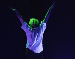 Jack, a white non-binary person with bright green hair and wearing a white t-shirt stands with their back to the camera under UV light, doing a morning stretch.