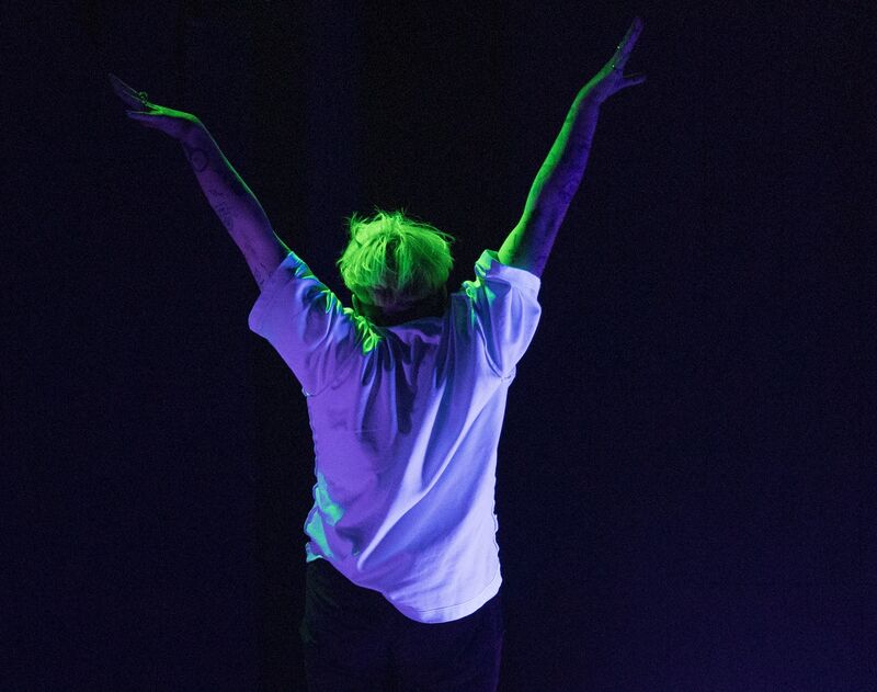 Jack, a white non-binary person with bright green hair and wearing a white t-shirt stands with their back to the camera under UV light, doing a morning stretch.