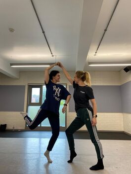 Two dancers dressed in dark colours, one has both legs on the floor and supports the other, who is balancing on one leg. They look at each other.