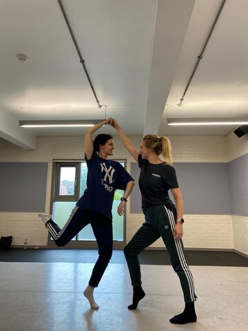 Two dancers dressed in dark colours, one has both legs on the floor and supports the other, who is balancing on one leg. They look at each other.