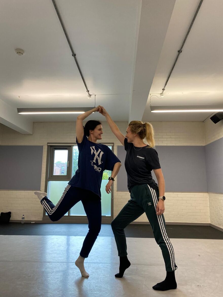 Two dancers dressed in dark colours, one has both legs on the floor and supports the other, who is balancing on one leg. They look at each other.