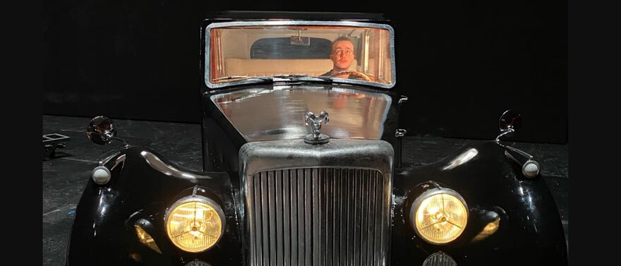Orion sitting in a model car on stage, the car's headlights lit up and light inside the cab of the car to make the actors visible. He is a white man with a small beard and moustache and dark hair tied back.