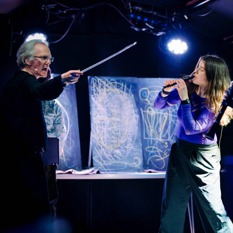 David, a white man with grey hair dressed in all in black, brandishes a white cane, mouth wide open as he barks instructions. Liza plays their recorder, body contorted to one side, controlled by his instructions. 