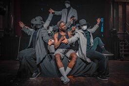 Sim Campbell sits centre stage on a sofa. He is a black man with dyed dreadlocks, wearing a black vest, grey shorts and white socks. He's surrounded by mysterious figures wearing trench coats, masks and hats. They frame him as they pass objects to eachother, behind and around him. But Sim is looking intently at his own hands, which are held up in front of him as he uses sign language.
