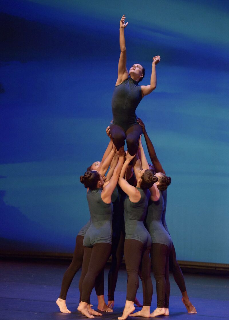 A group of dancers lifts a woman into the air while she reaches up with one arm.
