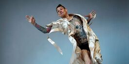 Male presenting figure, the artist Dan Daw, in mid-action pose in mid-air with arms outstretched. There is a mattress covered with a loose sheet beneath them. They are wearing a silk dressing gown that is open, exposing his tattooed skin.