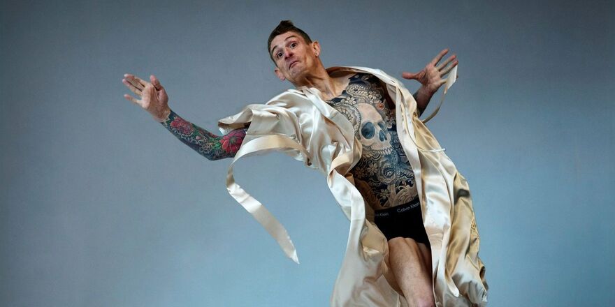 Male presenting figure, the artist Dan Daw, in mid-action pose in mid-air with arms outstretched. There is a mattress covered with a loose sheet beneath them. They are wearing a silk dressing gown that is open, exposing his tattooed skin.