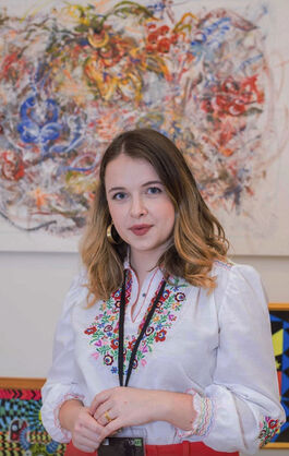 Photo of Ksenia Kazintseva in embroidered floral shirt in front of her abstract painting featuring floral patterns in blue and red.