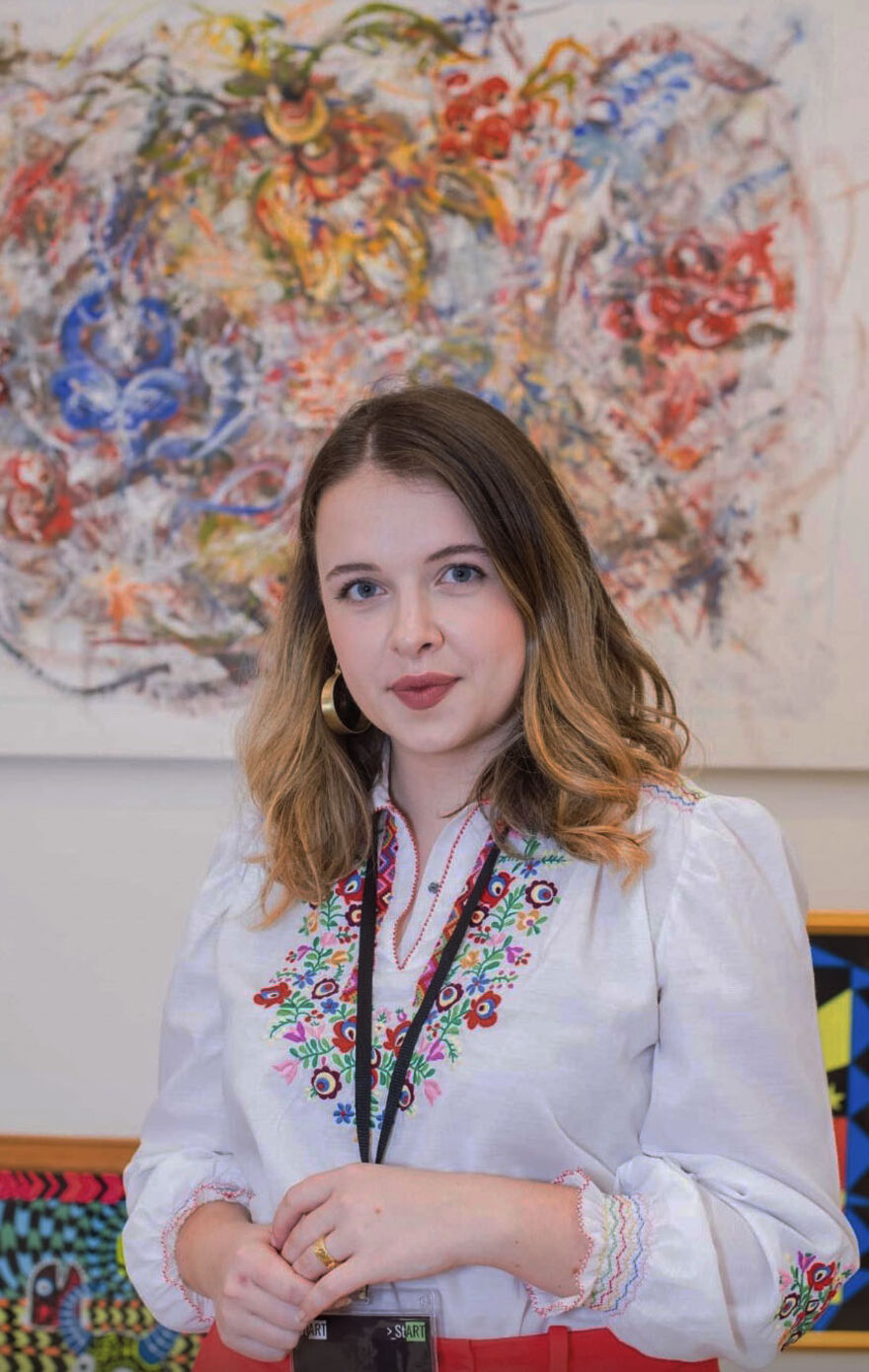 Photo of Ksenia Kazintseva in embroidered floral shirt in front of her abstract painting featuring floral patterns in blue and red.