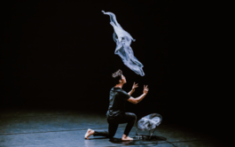 A man with short black hair and glasses is has their back slightly turned away from the viewer, wearing a black t-shirt. He has just thrown a pale, almost transparent piece of cloth in the air, his arms still outstretched from the motion. The cloth is still suspended in the air, and there is a fan in front of the man keeping it afloat. The background is completely black.