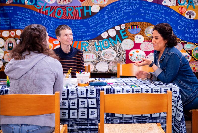 3 people sit around a table talking. It has a blue table cloth with white squares printed on it in front of a background of collaged art with lots of colours and words including 'Wandsworth'.