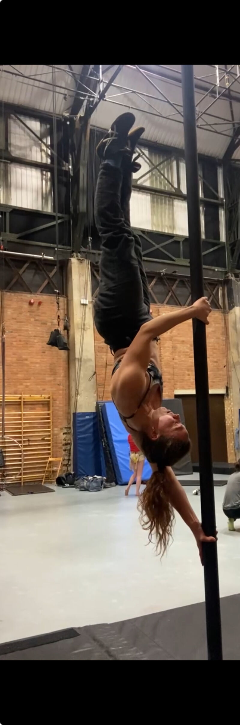 A woman hold herself upside down on a pole, with just her arms, in a flag, with her body pointing up to the ceiling .
