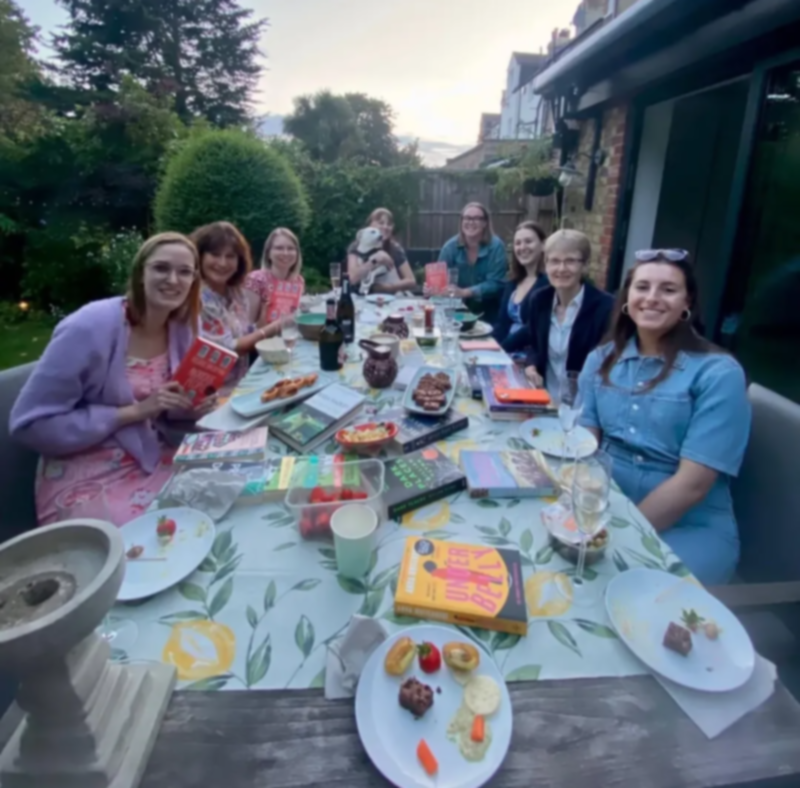 A diverse group of women meeting together in the sunshine to discuss a book with snacks.