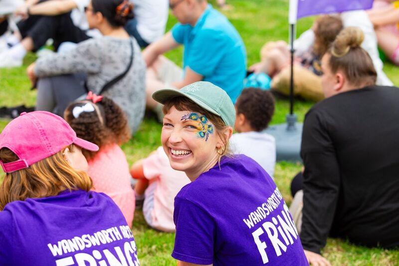 In A Crowd Of People Sitting On Grass, A Person With An Elegant Facepaint Design Over Their Eyebrow And Cheek Is Turning To The Camera And Smiling As They Look Directly At You. They Are Wearing A Peaked Cap And Purple T-shirt Bearing The Words Wandsworth Arts Fringe.