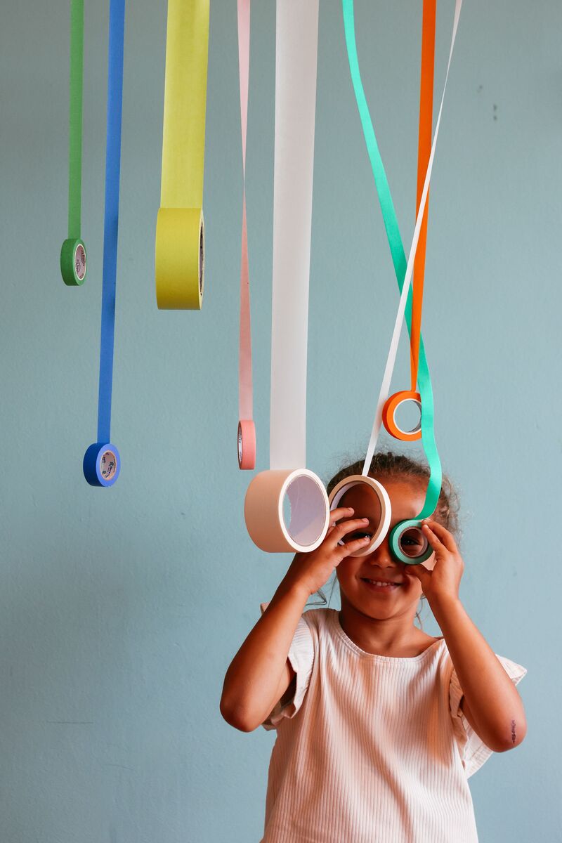 A young girl is standing beneath tape hanging from the ceiling. She is peeping through the holes in the rolls of tape.