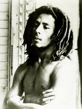 A portrait of Bob Marley taken in 1977. He is topless and wears his hair down and his arms folded, though he looks relaxed.