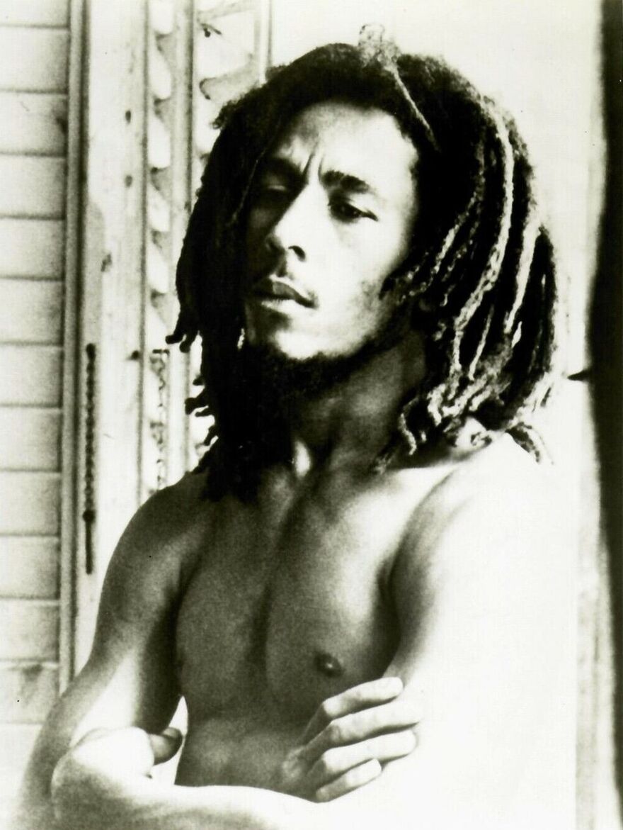 A portrait of Bob Marley taken in 1977. He is topless and wears his hair down and his arms folded, though he looks relaxed.
