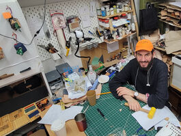 Shows the inside of a creative workshop with a bearded man with an Orange hat sitting at his workbench with a yellow mallet. Surrounded by printers, hand tools and paints.